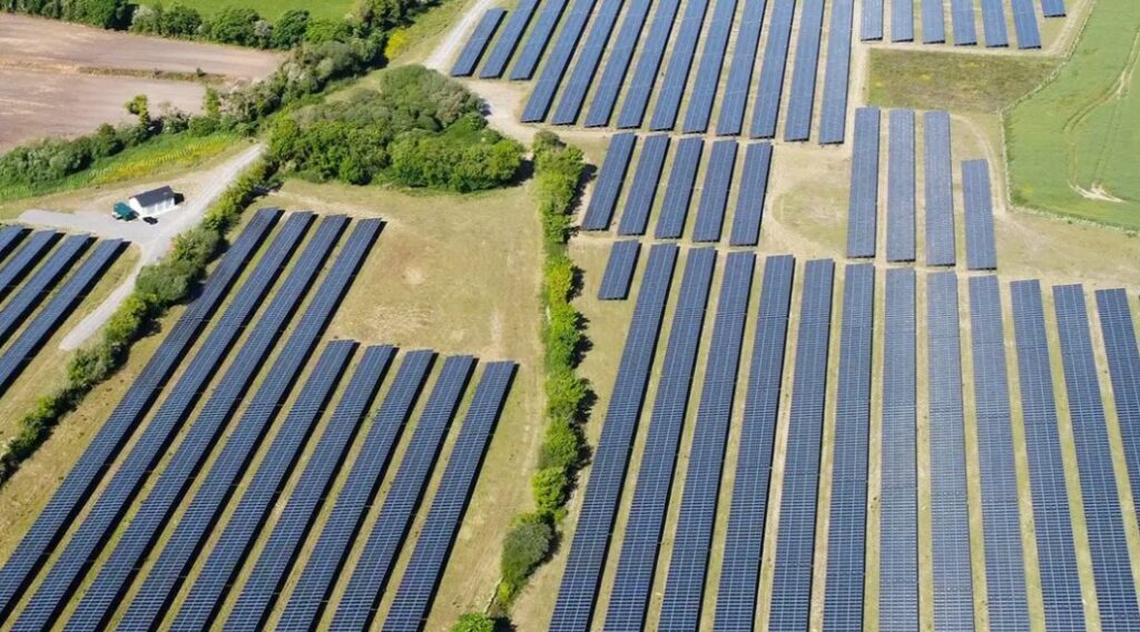 a field of solar panels from above with a small cottage in the background