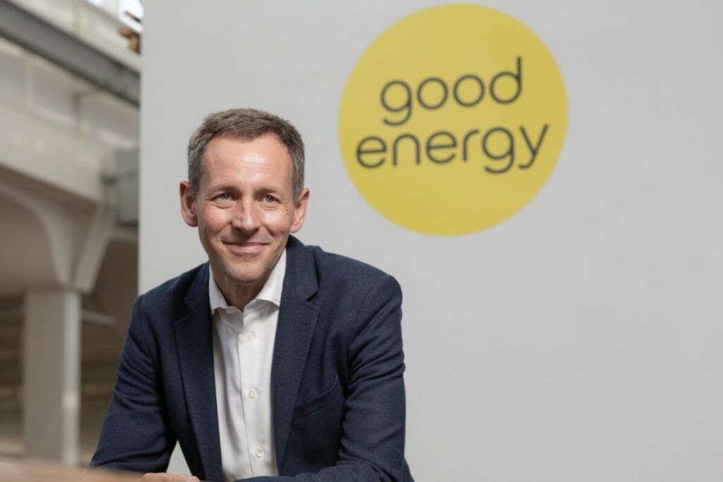 a white man in a suit gives a small smile as he sits in front of a wall with the good energy logo