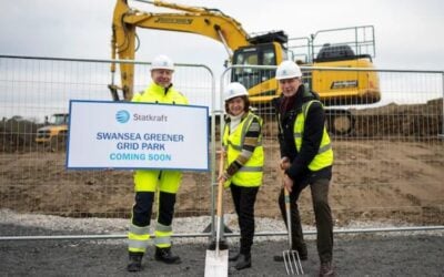 Three people stand in front of a large digger holding a sign announcing the greener grid project, spades, and pitchforks.