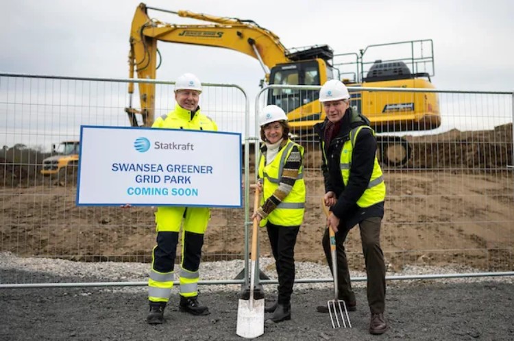 Three people stand in front of a large digger holding a sign announcing the greener grid project, spades, and pitchforks.